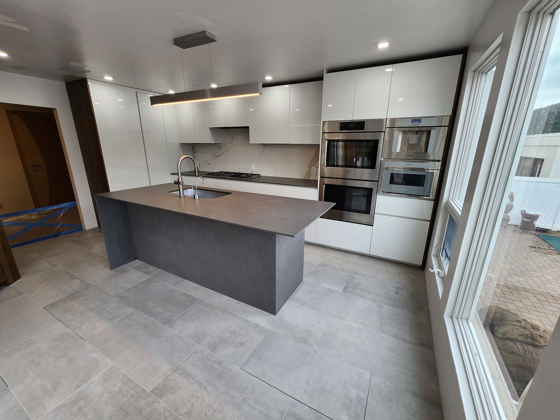 Modern kitchen with white cabinetry, stainless steel appliances, and a gray island. Large windows provide natural light.