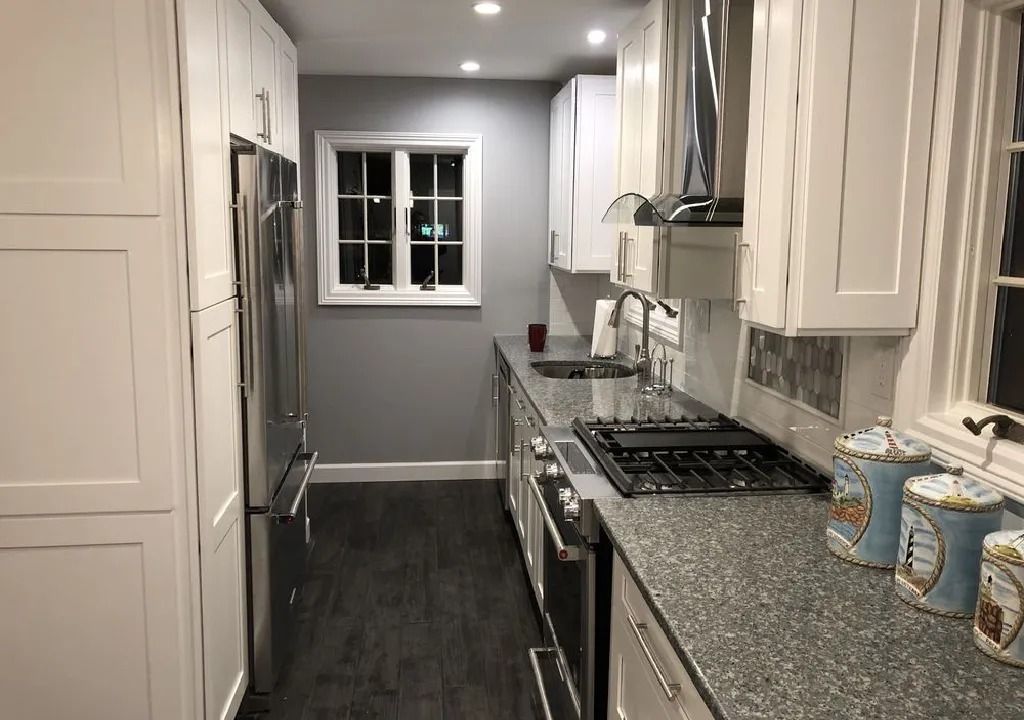 A narrow kitchen with white cabinets, stainless steel appliances, gray countertops, and a dark wood floor. A small window is centered on the gray wall.