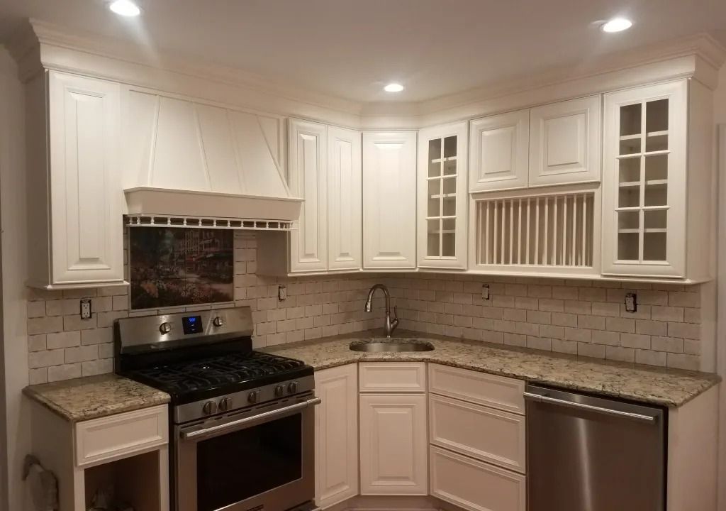 White kitchen with granite countertops, stainless steel appliances, and light-colored backsplash tiles. Upper cabinets line the walls, and a corner sink sits in the middle.