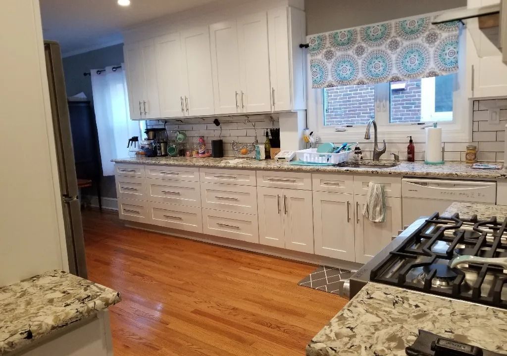 A kitchen with white cabinets, granite countertops, and wood floors. A gas stove is in the foreground, and a window with patterned curtains is visible.