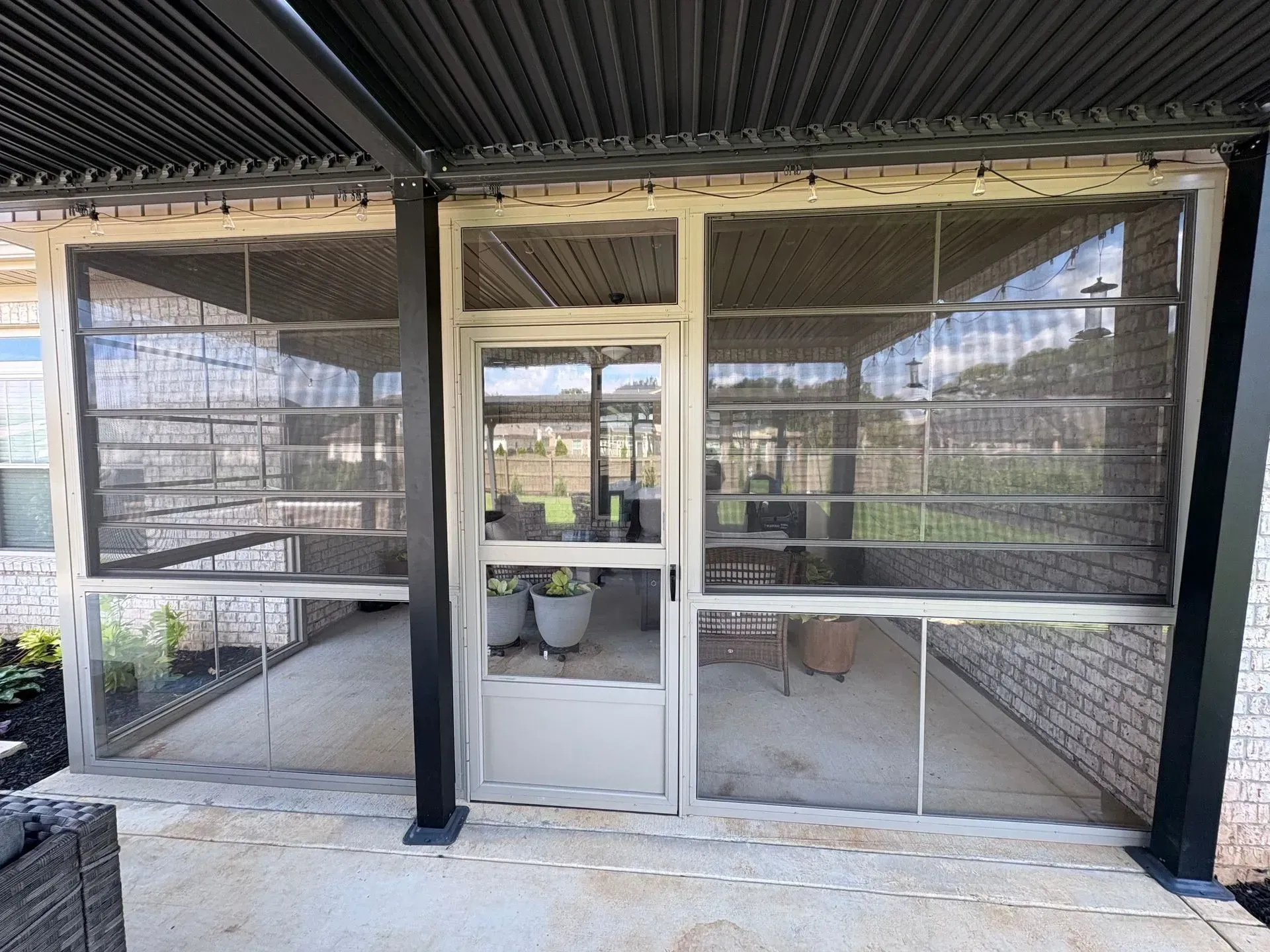 Screened-in patio with door. Beige and black brick and frame. Plants and outdoor furniture visible inside.