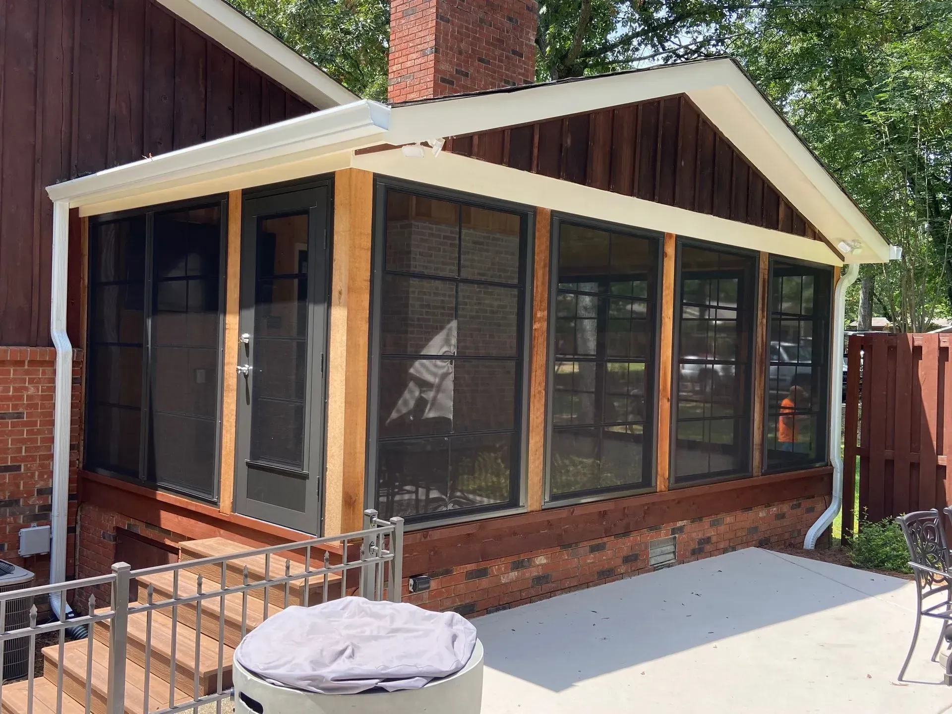 Screened porch with dark screens, wooden frames, and brick base attached to a brown-sided house.