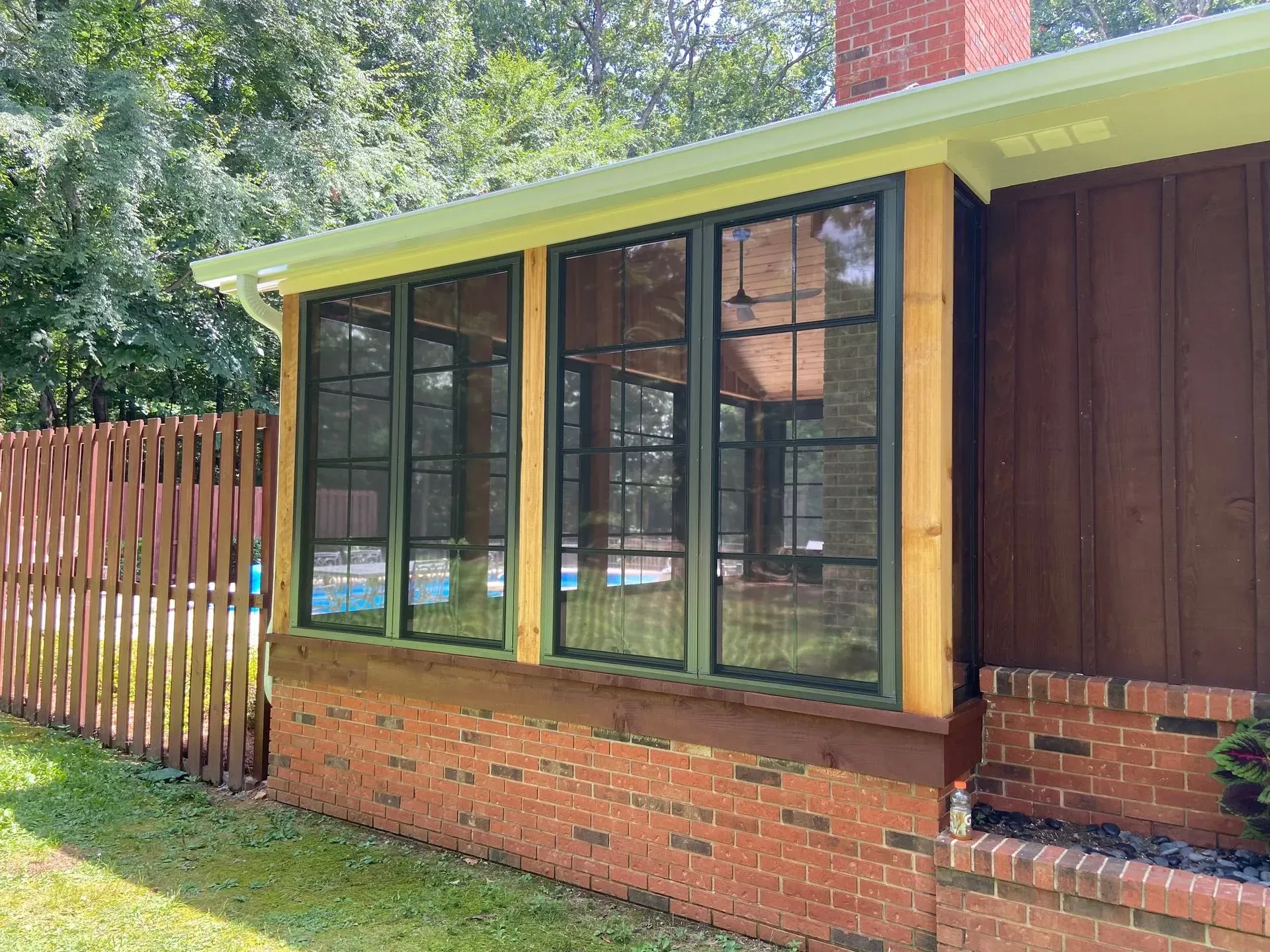Screened-in porch with green frames, brown siding, brick base, and wooden fence in a grassy yard.