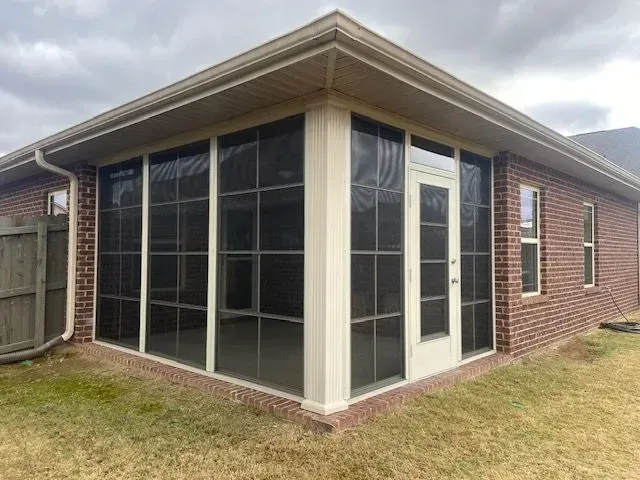 Screened-in porch attached to a brick house. Cream-colored frames with dark screens. Overcast sky.