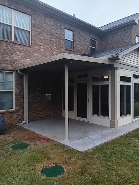 Covered patio next to a brick house with windows, adjacent to a sunroom on the right, green grass.