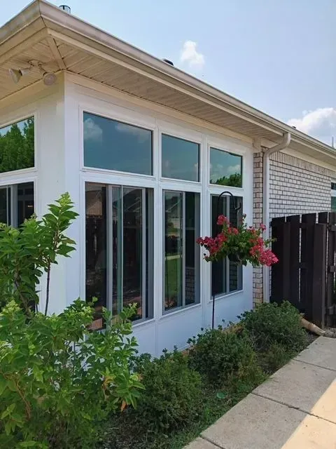 White-walled sunroom with large windows, hanging flower basket, and lush green bushes. Light-colored brick wall in the background.