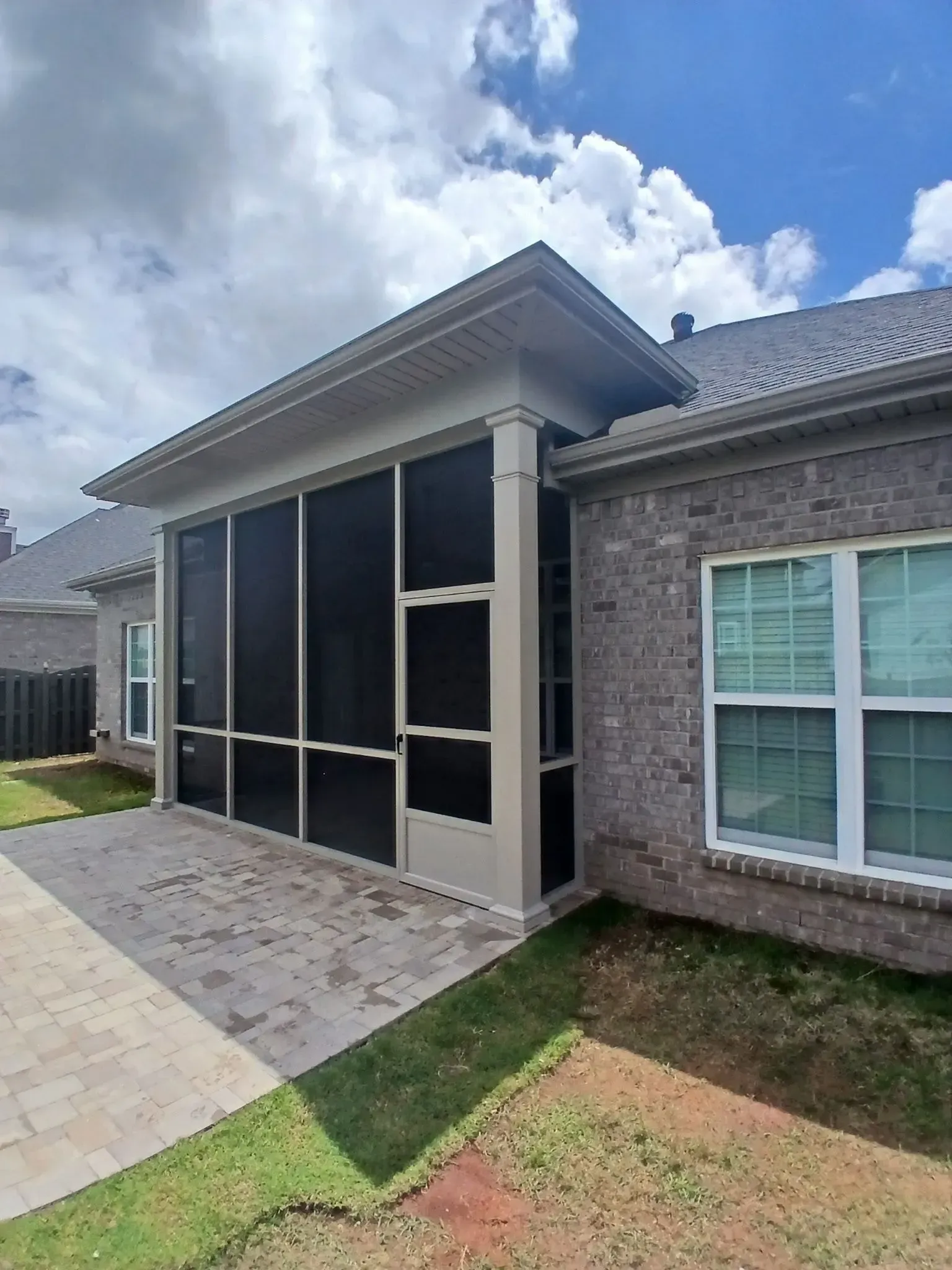 Screened-in porch with beige supports, brick house, and stone patio on a sunny day.