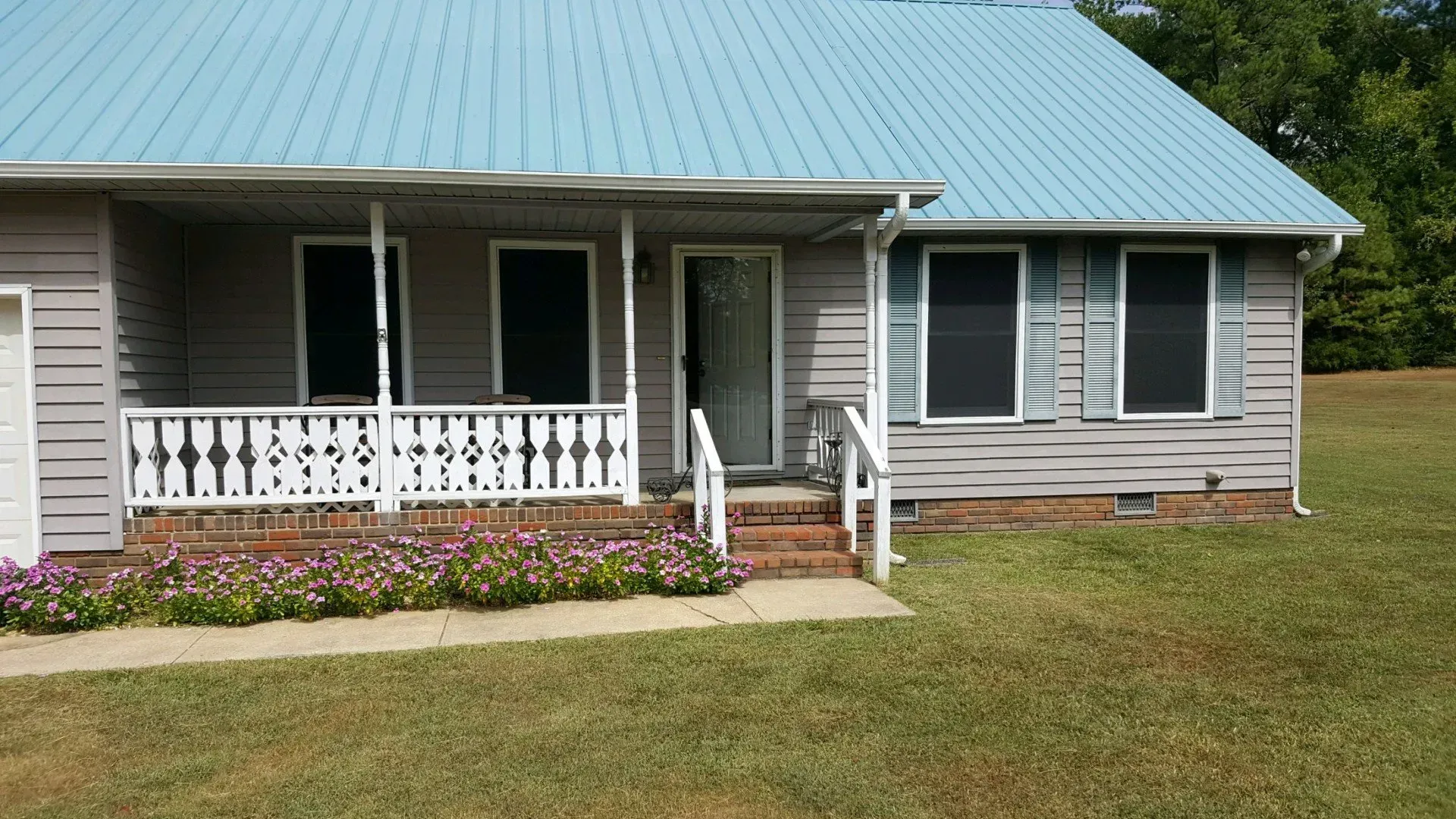 A house with a blue metal roof, front porch, and yard with flowers and grass.