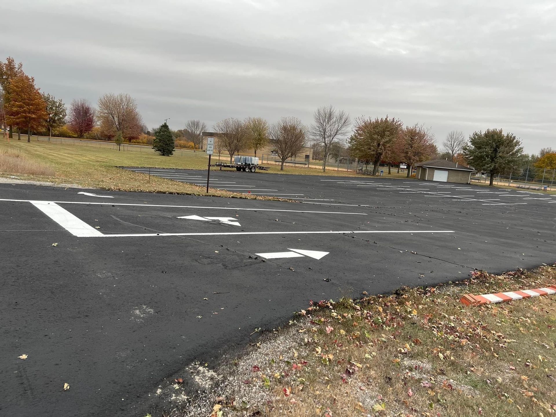 Newly paved parking lot with directional arrows, next to grassy area with trees and cloudy sky.