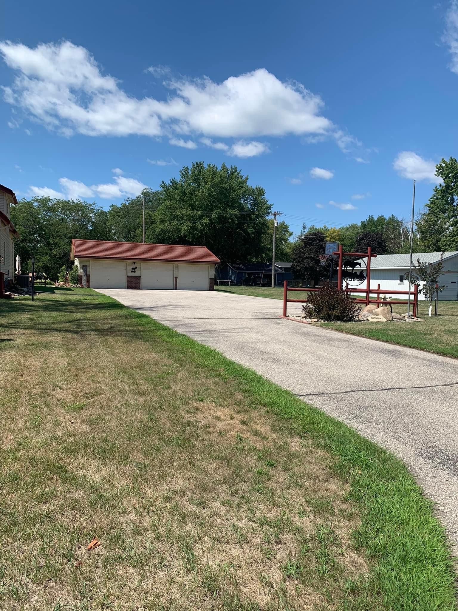 Concrete driveway leads to a small building with a red roof, basketball hoop on right, blue sky with clouds.
