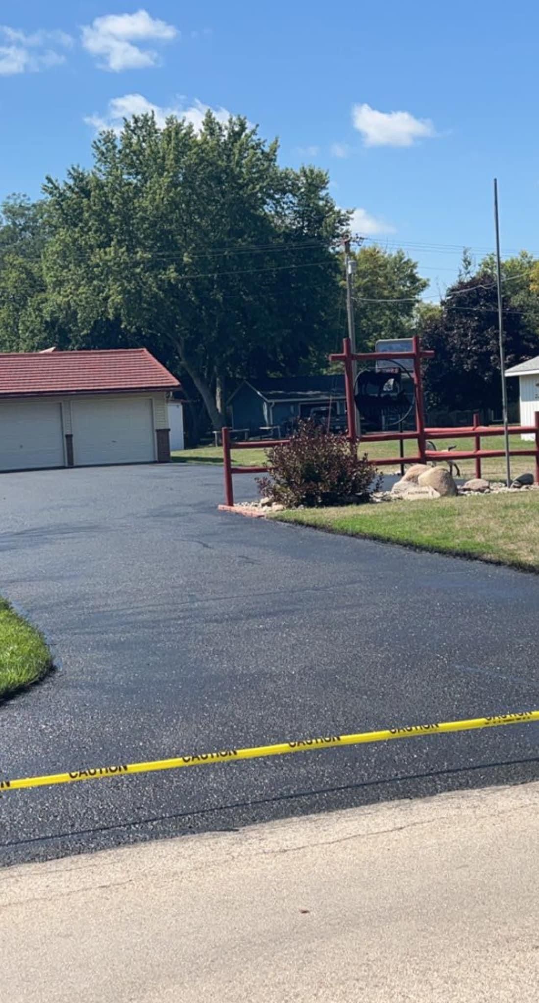 Newly paved black driveway with caution tape, grass, and buildings under a blue sky.