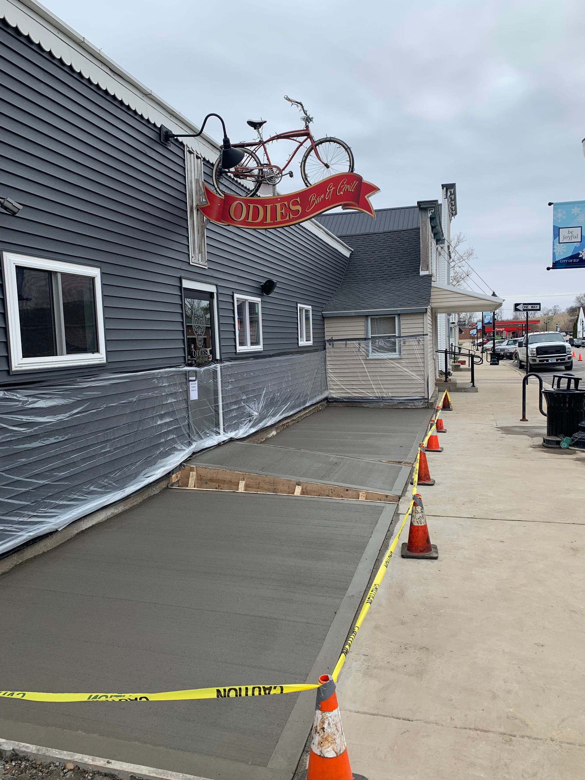 New concrete sidewalk in front of a gray building with bike sign, yellow caution tape, and orange cones.
