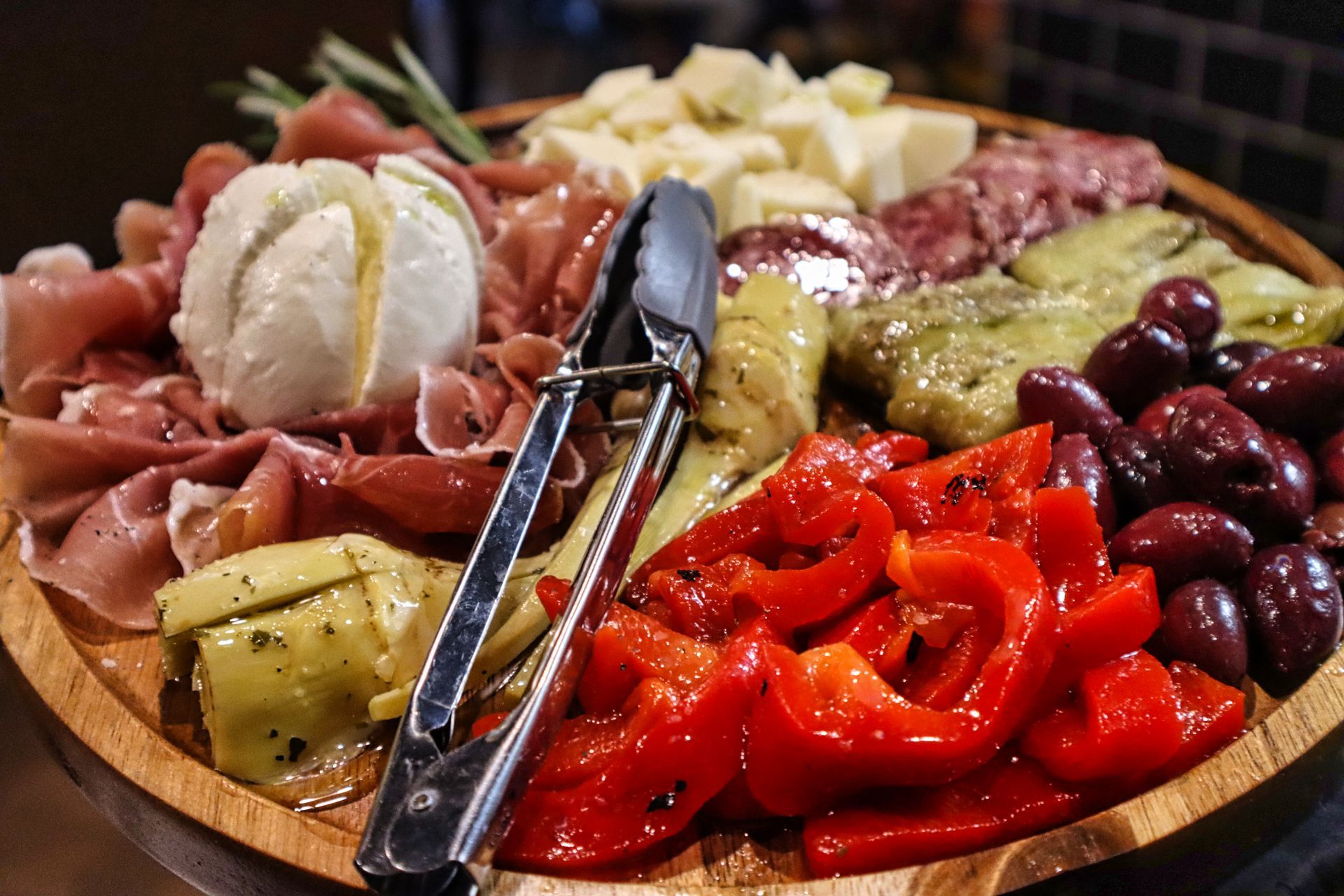 An antipasto platter on a wooden board featuring prosciutto, burrata, marinated artichokes, roasted peppers, and olives.