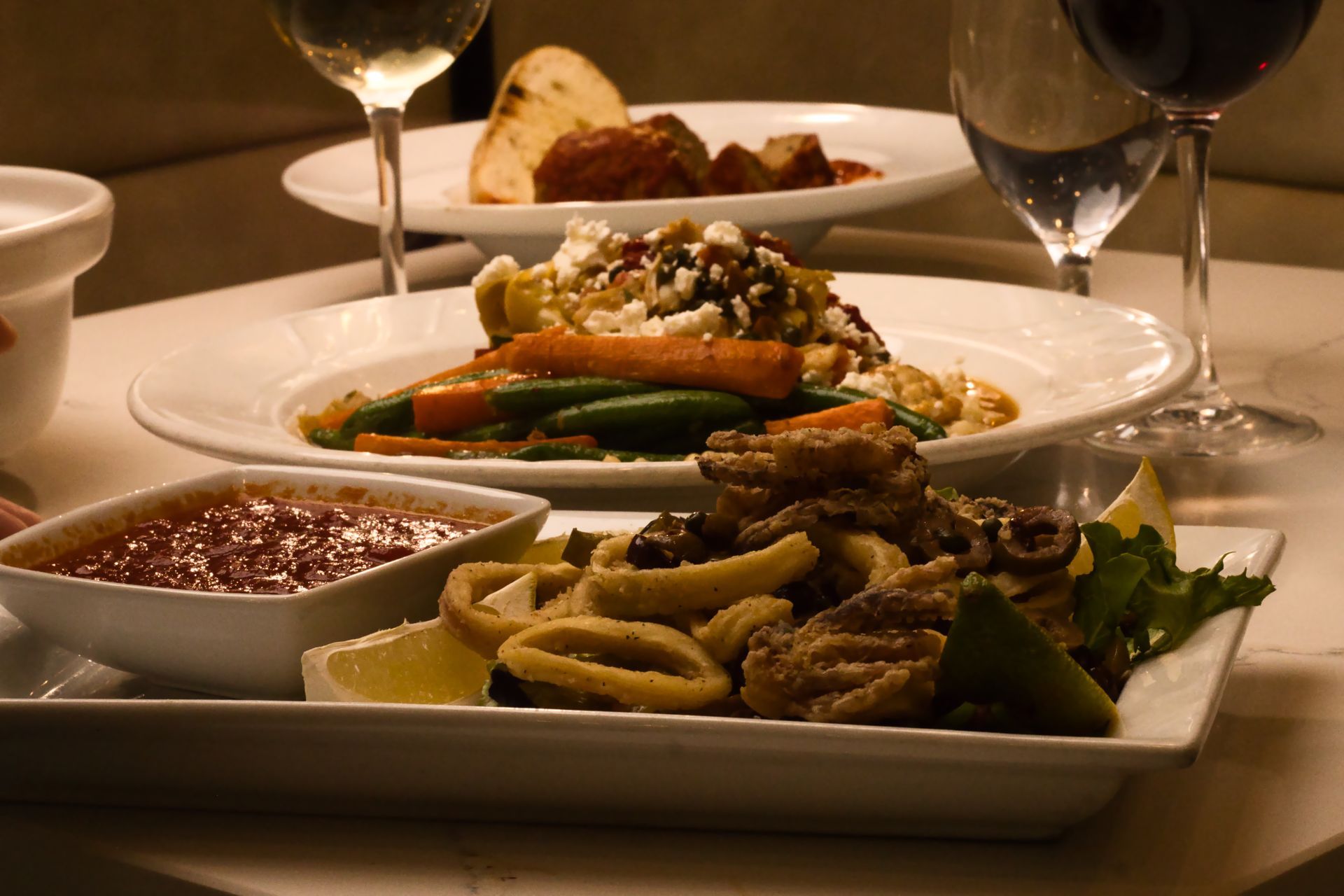 A table setting with several plates of food, including fried calamari, vegetables with cheese, and wine glasses.