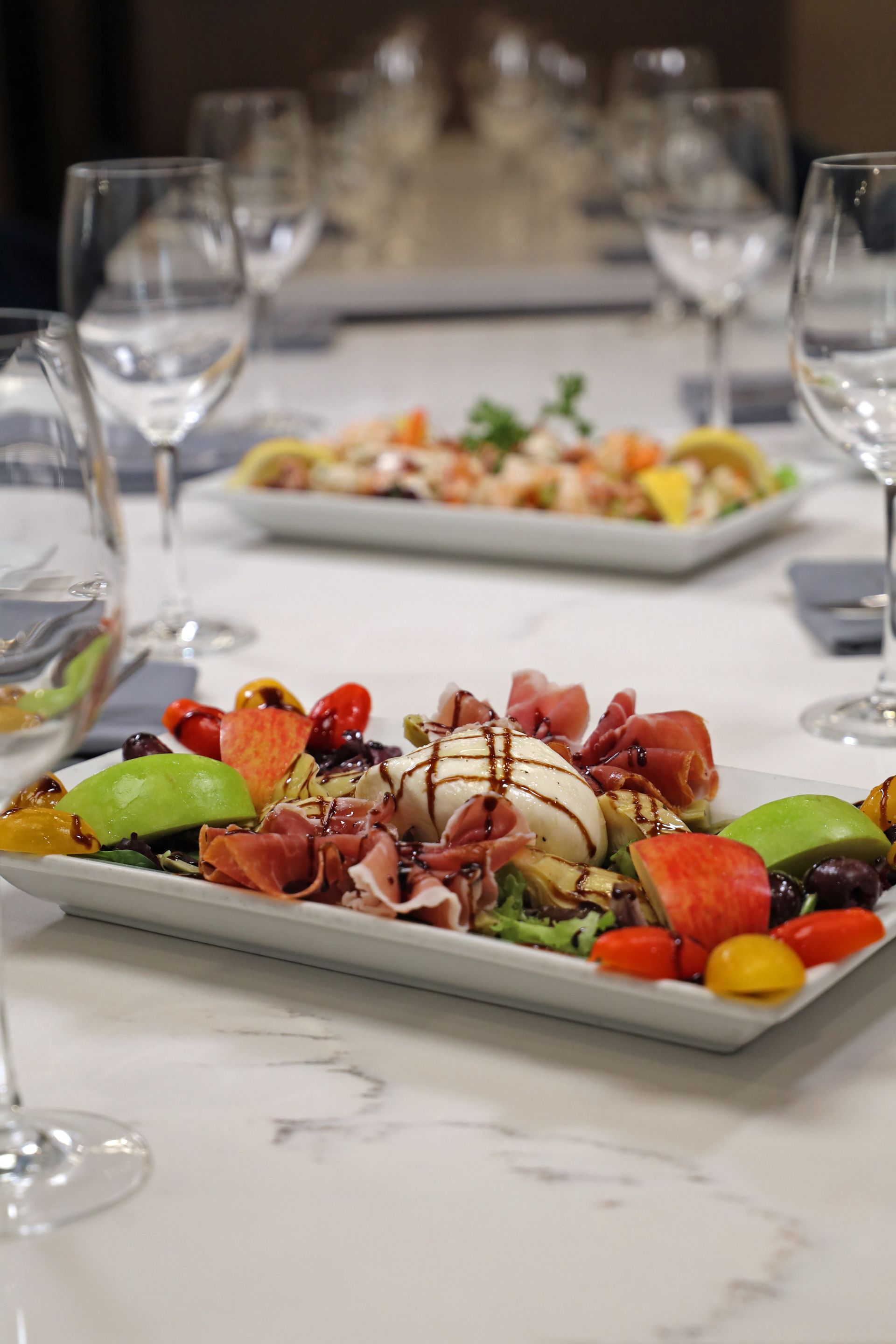 Two rectangular plates of appetizers featuring burrata, prosciutto, and fruit, served on a table with wine glasses.