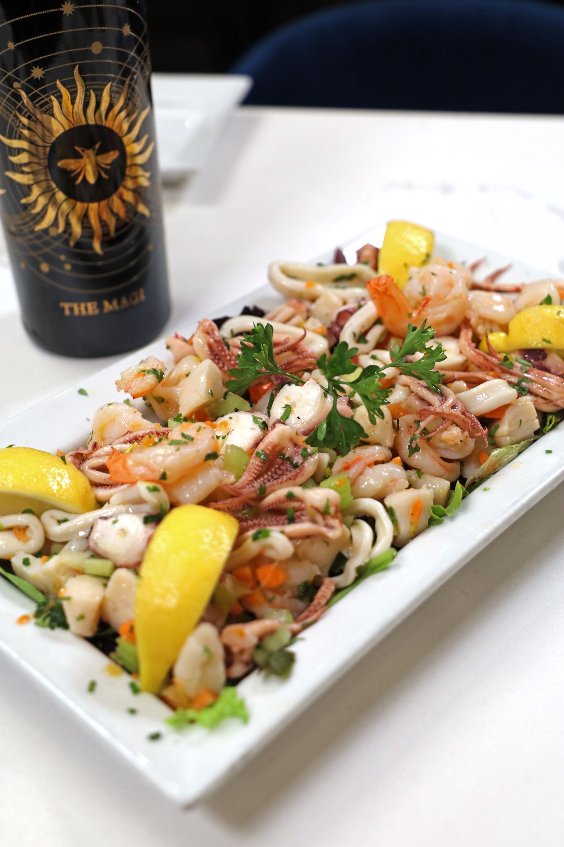 A platter of seafood salad garnished with lemon wedges and fresh parsley, set next to a wine bottle on a white table.
