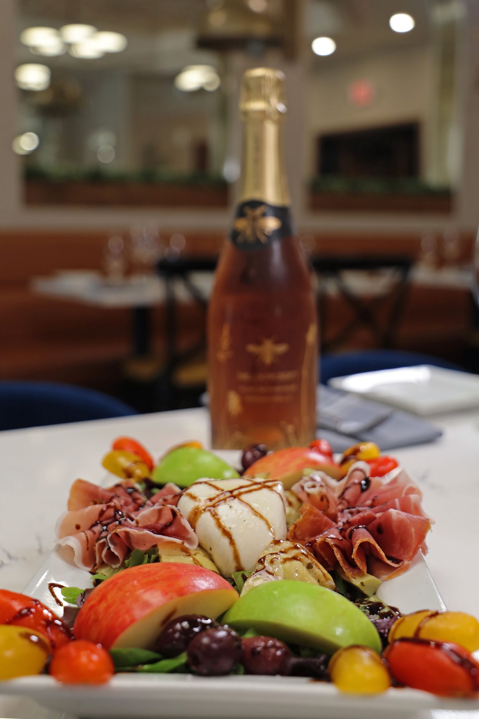 A plate of charcuterie, burrata, and fresh fruit sits in the foreground with a bottle of sparkling wine behind it.