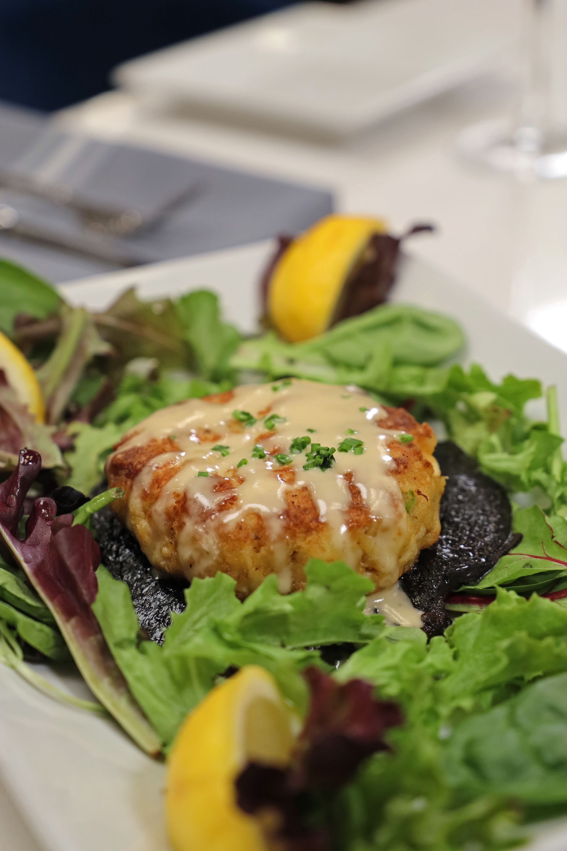 A crab cake topped with sauce, served on a bed of greens with lemon wedges on a white plate.