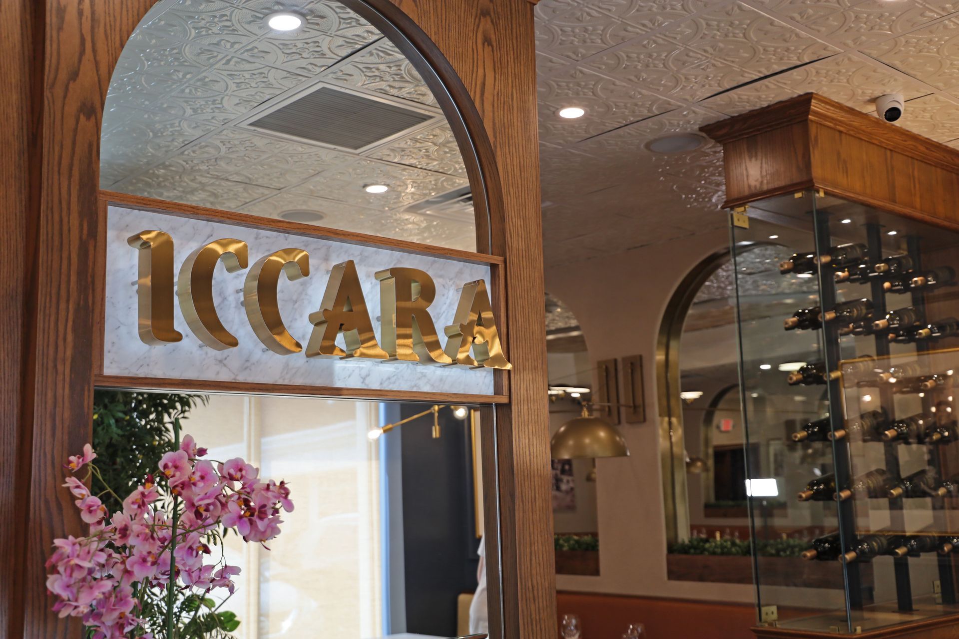 The gold-lettered sign for Iccara hangs above a restaurant entrance, accented by pink orchids and a glass wine rack.