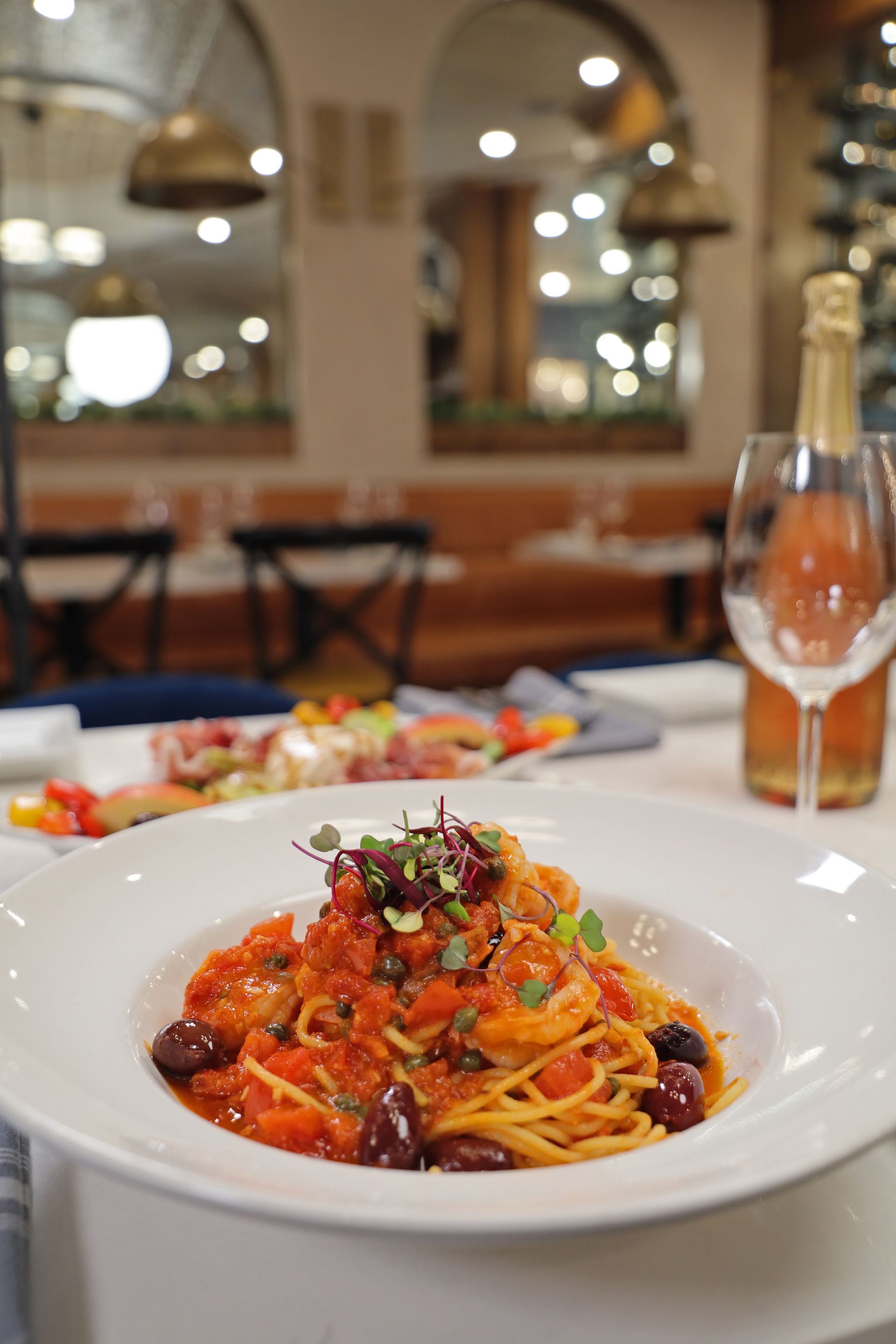 A bowl of tomato-based pasta with olives and shrimp sits on a restaurant table next to a glass of wine and another dish.