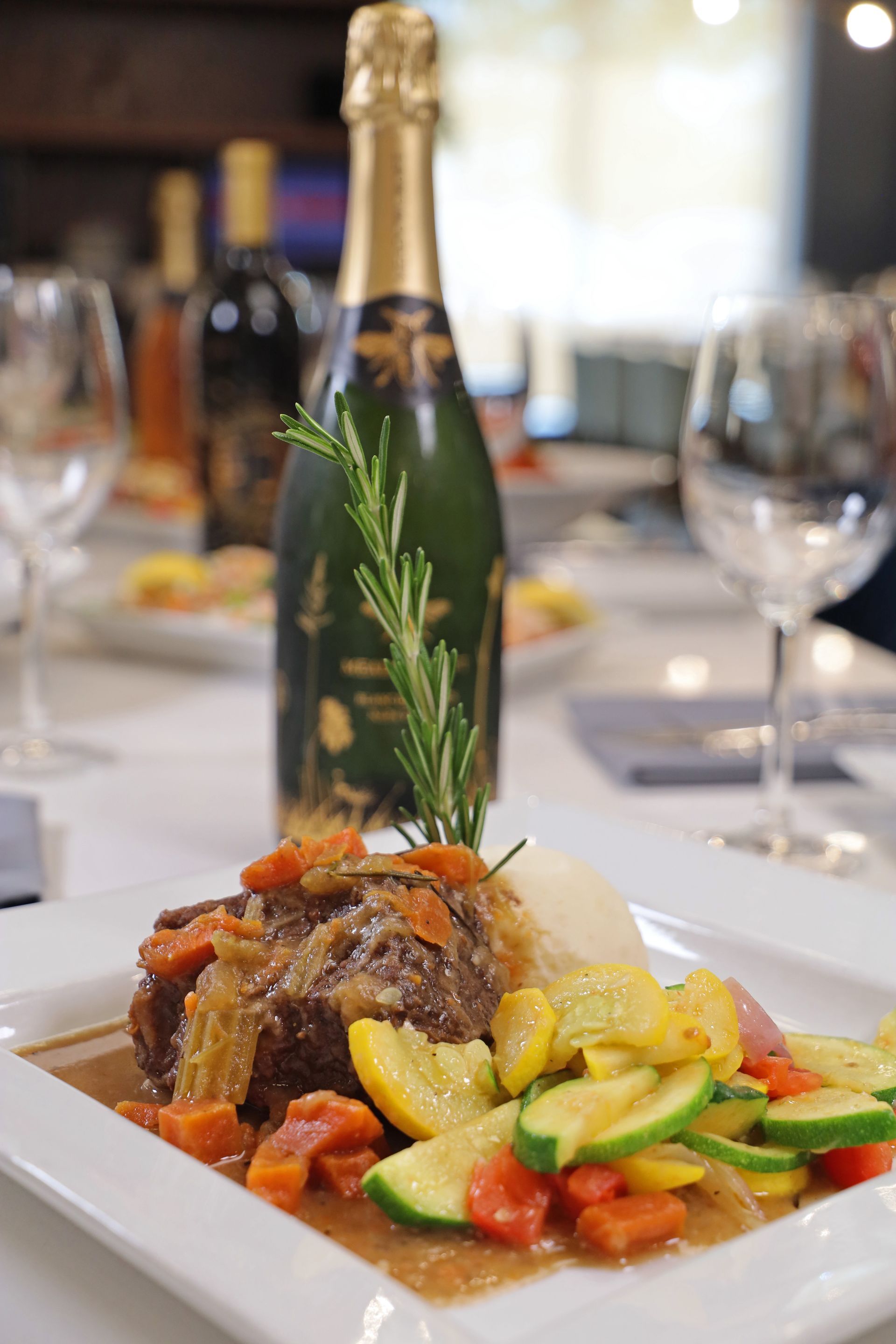 A plated meal featuring braised meat, carrots, and squash, garnished with rosemary, set before a wine bottle and glasses.
