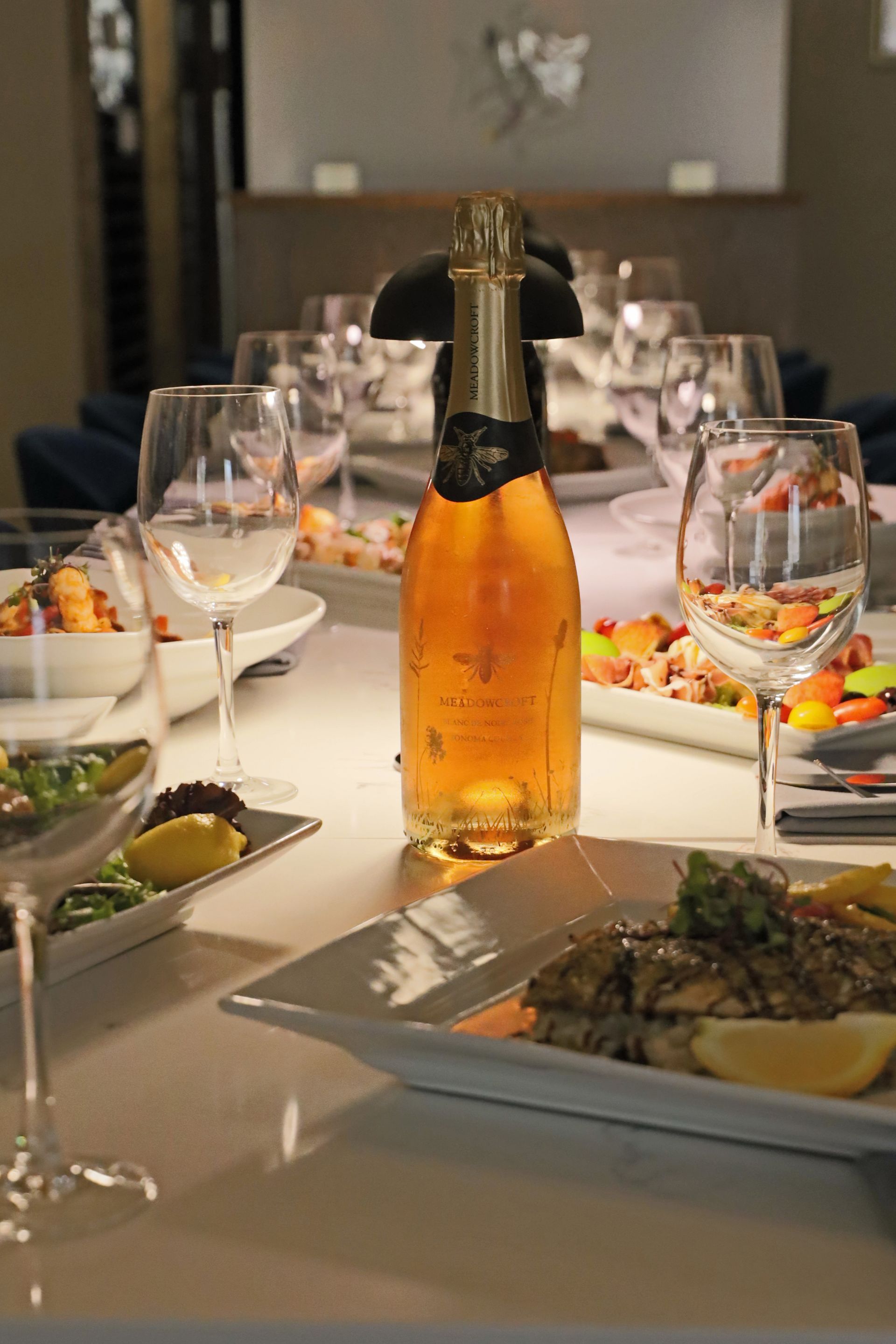 A table set for a fine dining meal with a central bottle of champagne, wine glasses, and plates of gourmet food.