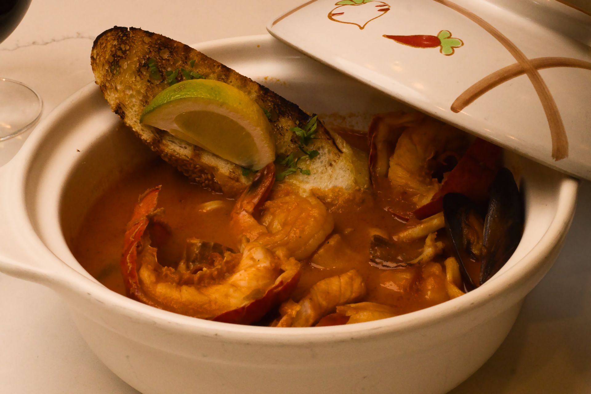 A white ceramic pot filled with seafood stew, garnished with a toasted bread slice and a lime wedge, partially covered.