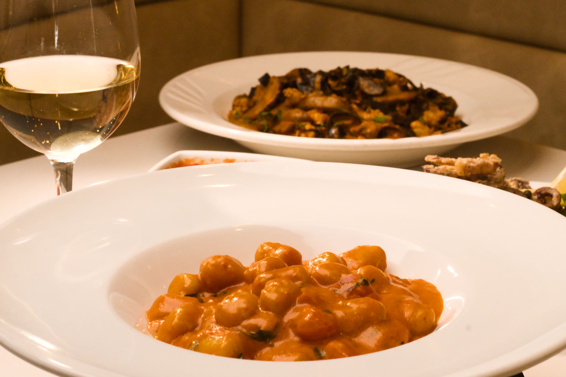 A white bowl of gnocchi in pink sauce sits in the foreground with a dish of mushrooms and a glass of white wine nearby.