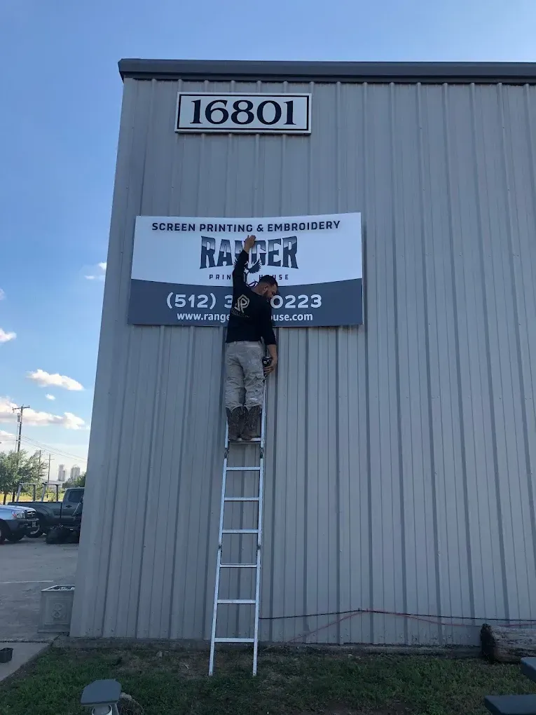 Person on a ladder attaching a sign to a gray building under the number 16801.