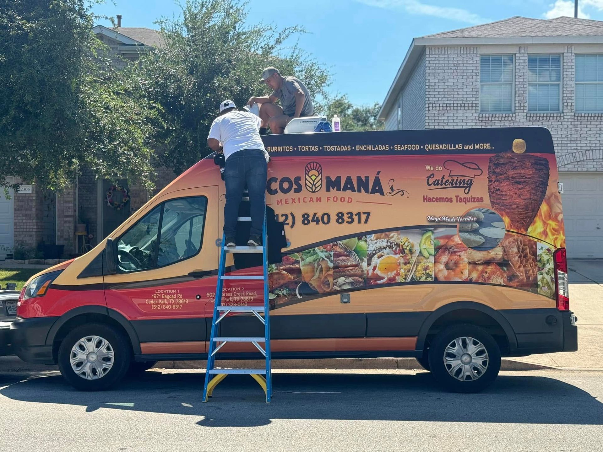 Red food truck with colorful menu wrap parked on a street while two workers stand on a ladder cleaning the roof.