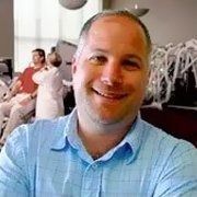 A smiling person with short brown hair wearing a light blue checkered shirt, with a blurred indoor scene in the background.
