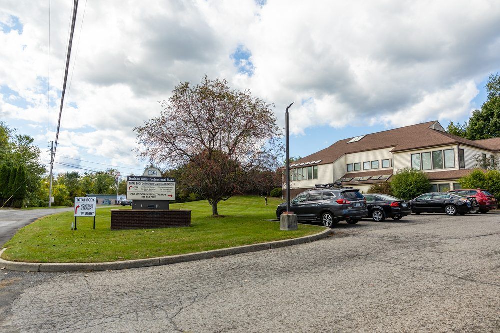 A two-story tan building with a brown roof, a lawn with a sign, and parked cars under a partly cloudy sky.