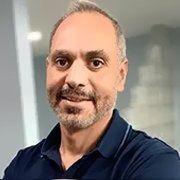A smiling man with a short beard, wearing a dark blue polo shirt, captured in a portrait shot against an office background.