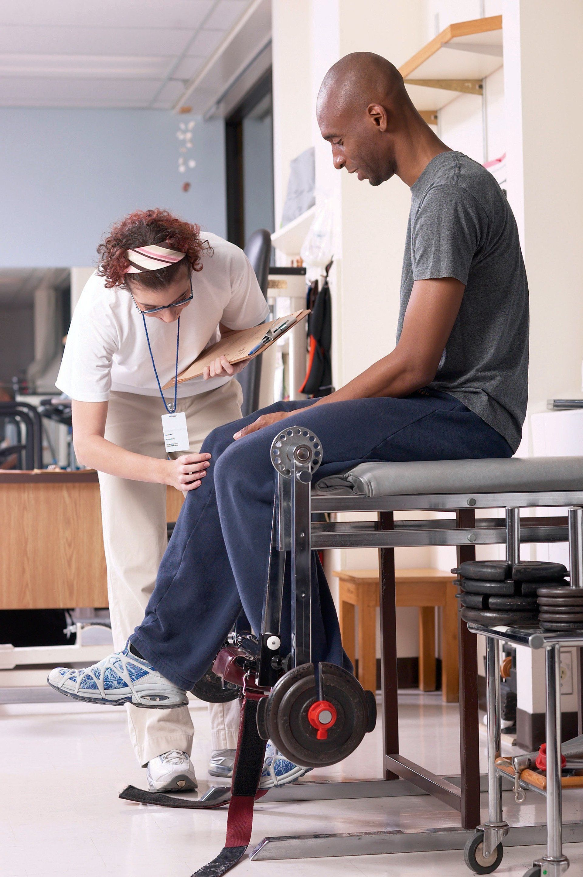 A physical therapist assists a patient with a leg exercise using a weighted machine in a clinical setting.