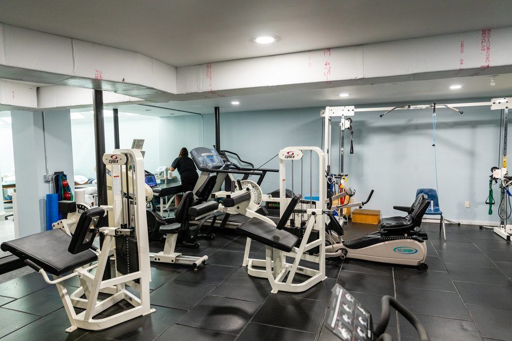 A well-lit basement gym with white walls, black flooring, various weightlifting equipment, and a stationary exercise bike.