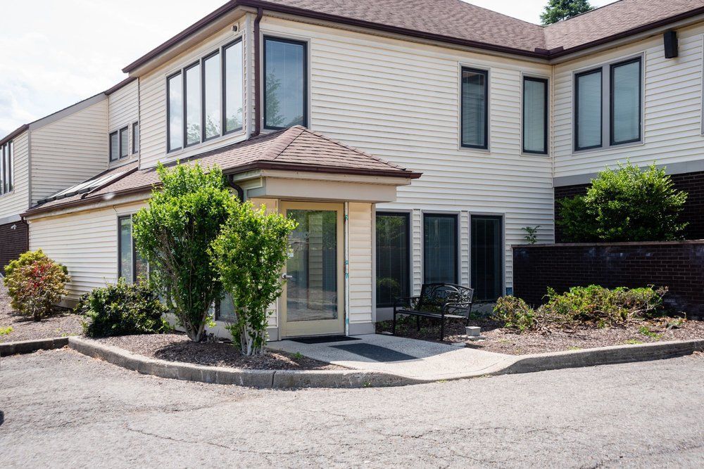 A beige two-story commercial building with a glass-paned entrance, surrounded by landscaped bushes and a parking area.