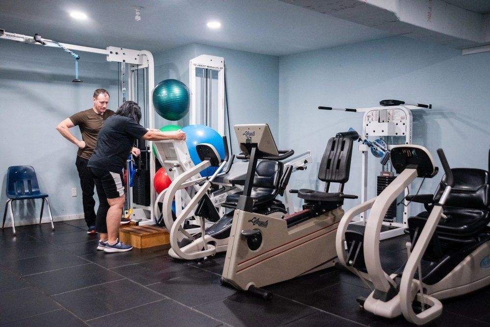 A physical therapist assists a person using exercise equipment in a brightly lit gym.