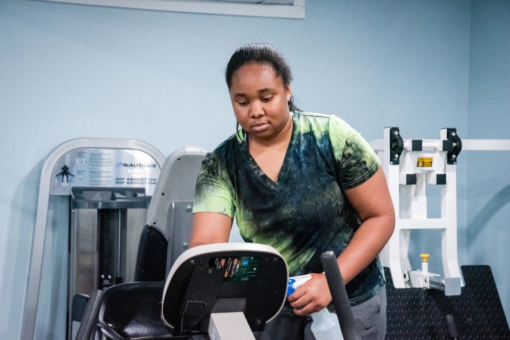A person in a green-and-black tie-dye shirt wiping down the console of a gym machine with a cleaning cloth.