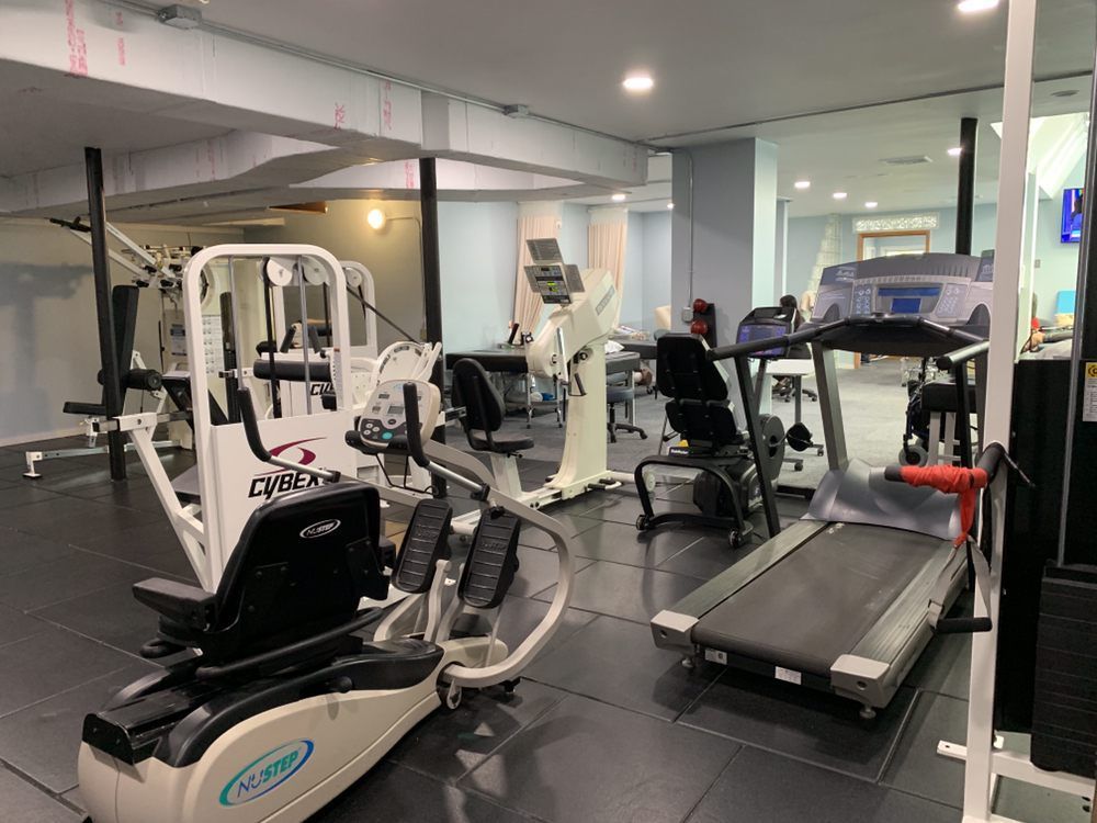 A well-lit indoor gym with various exercise machines, including a treadmill and stationary bike, on a black rubber floor.