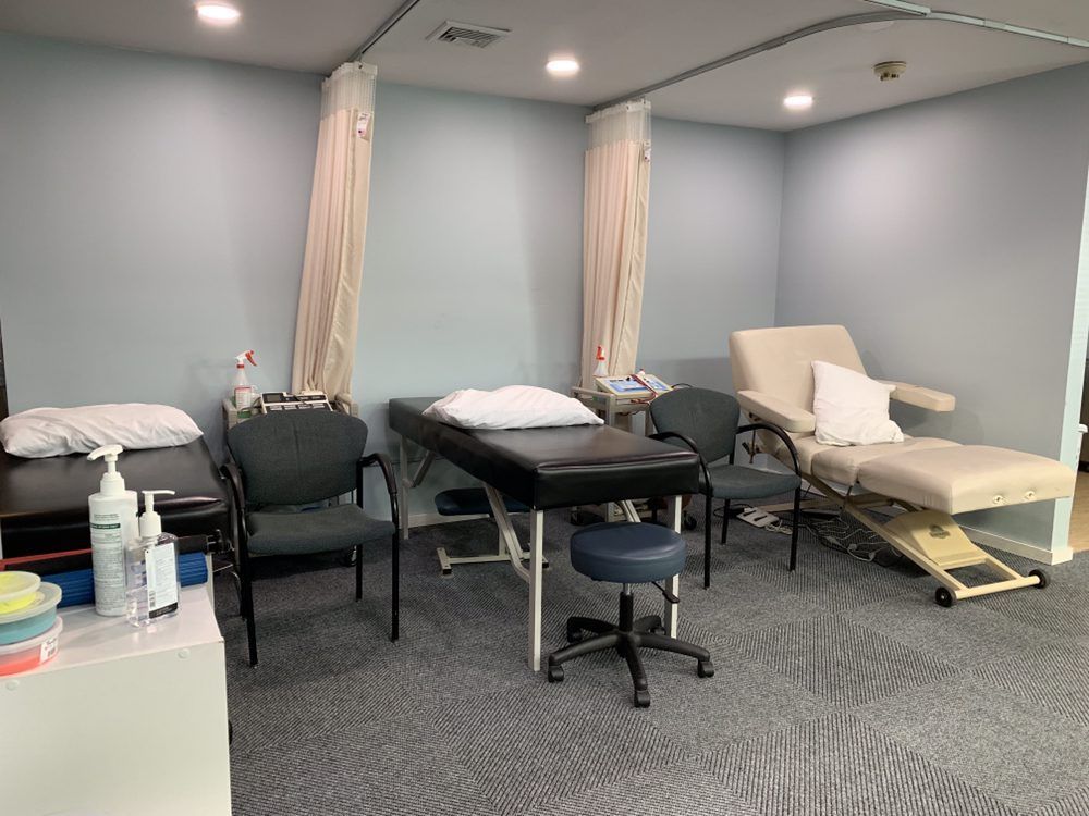 A medical treatment room featuring two black exam tables, a beige reclining chair, office chairs, and privacy curtains.