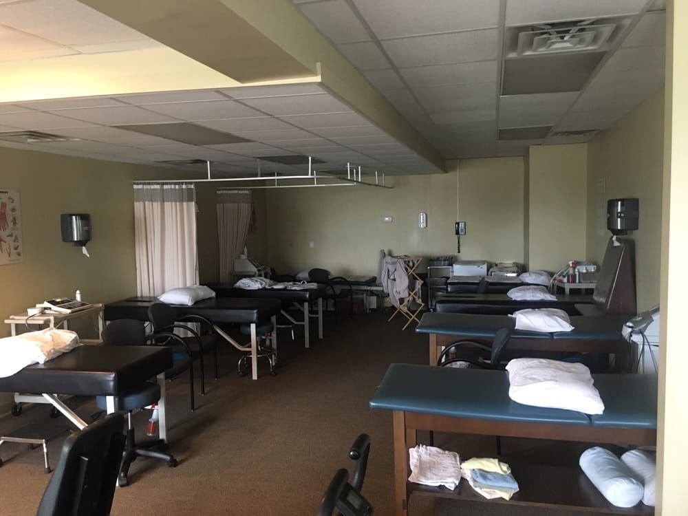 A physical therapy or clinical room with multiple massage tables, white pillows, privacy curtains, and rolling stools.