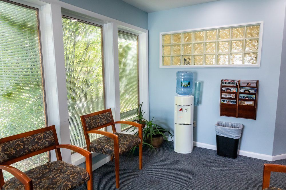 Waiting room with two patterned chairs, a water cooler, a brochure rack on a blue wall, and large windows with blinds.