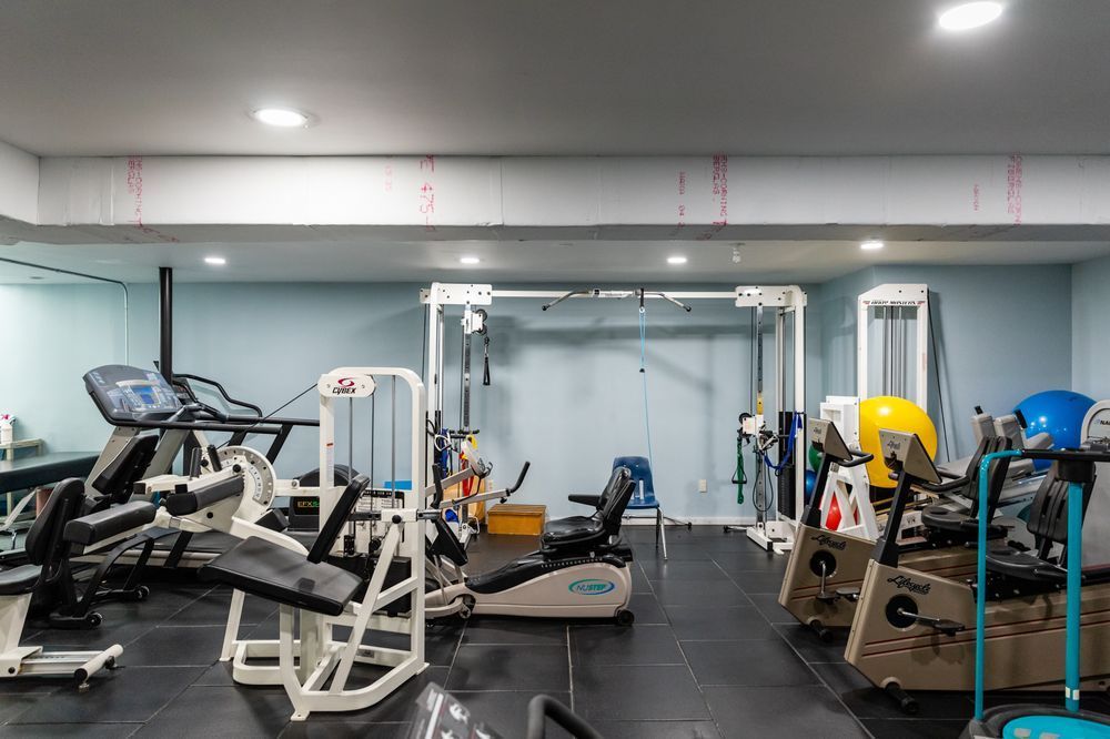 A home gym with various exercise machines, including a treadmill and weight stations, on a dark rubber floor.