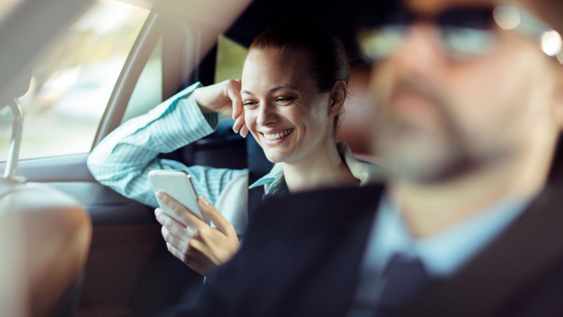 businesswoman in a professional blue shirt smiles while looking at her smartphone in the backseat
