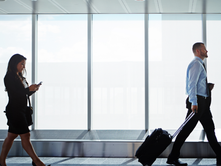 A smiling family of four is taking a selfie at an airport terminal, with a limousine and a chauffeur in the background. They're casually dressed for travel, standing next to their luggage, ready to depart. This scene suggests they might be using a limousine service, such as Rockford Rides, for comfortable transportation to Chicago O'Hare Airport.