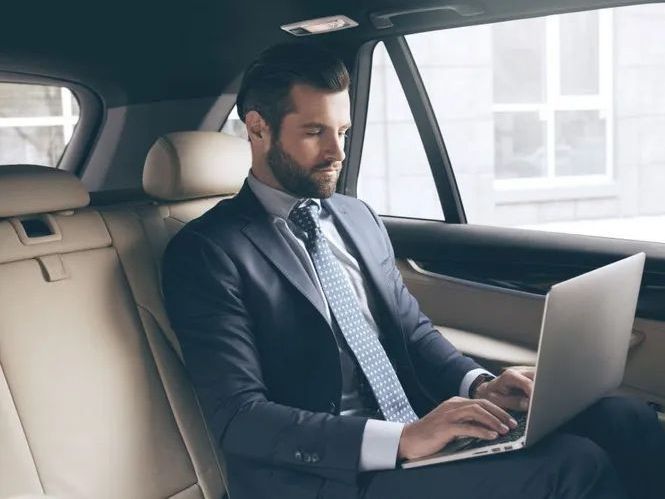 A man in a suit and tie is sitting in the back seat of a car using a laptop computer.