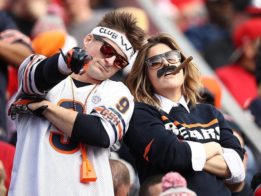 Two exuberant Chicago Bears fans are enjoying the game day atmosphere, dressed in team jerseys and sporting playful accessories like sunglasses, a headband, and fake mustaches. They embody the high spirits and camaraderie that fans can expect when traveling to a Bears game with Rockford Rides, which offers party bus services from Rockford to Chicago, ensuring fans arrive at Soldier Field ready to cheer on their team.