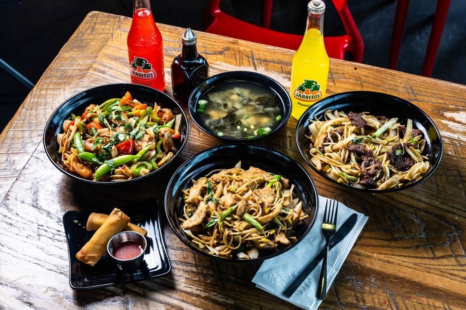 Table with Asian food bowls, soup, soda bottles, and silverware on a wooden table.
