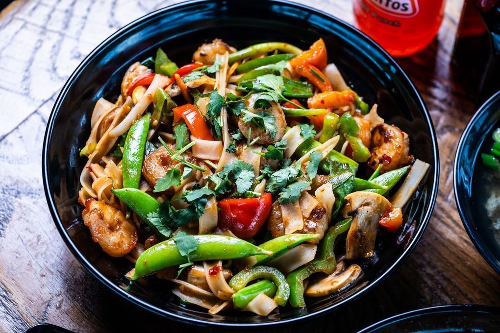 Black bowl of stir-fry noodles with vegetables, shrimp, and cilantro.