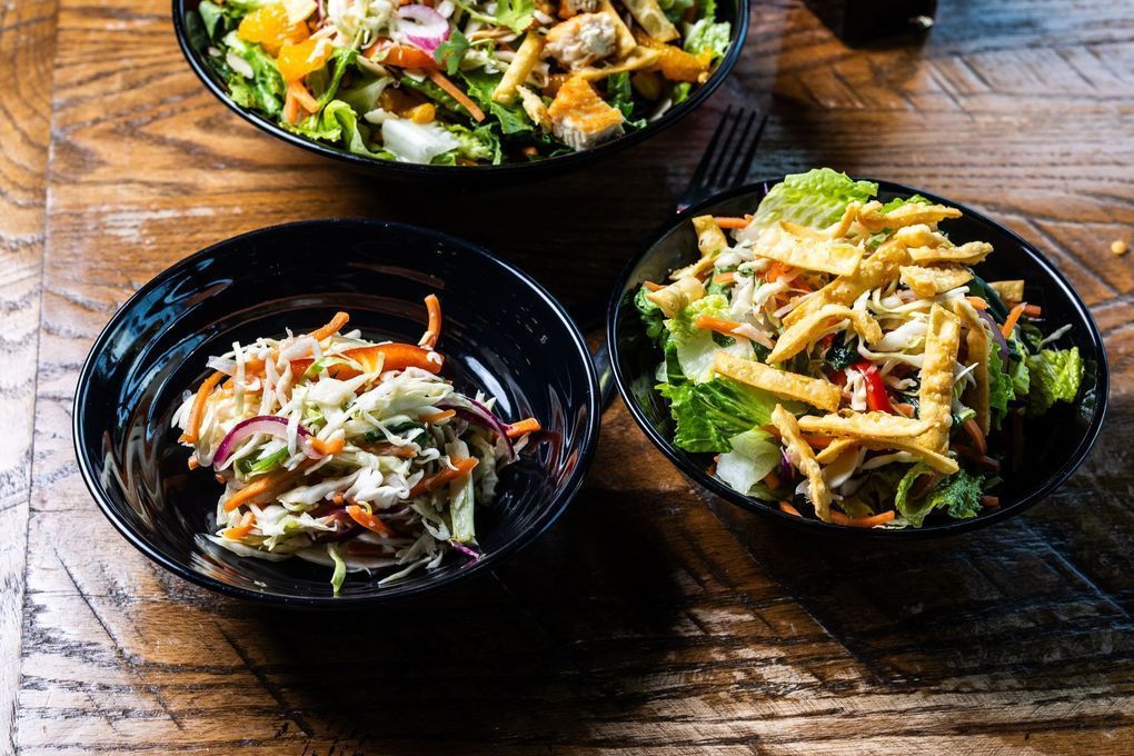 Three salads in black bowls on a wooden table. The salads have various toppings and colors.